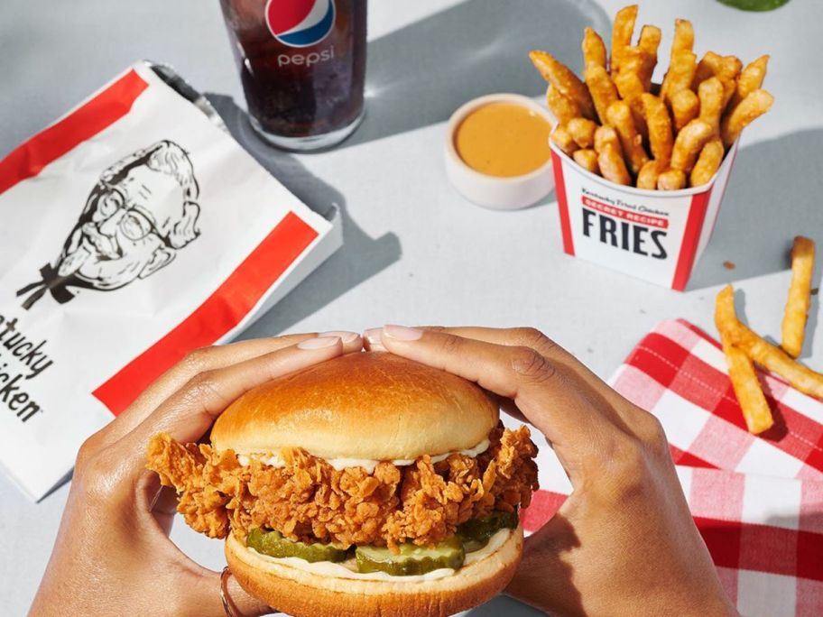 Hands eating a KFC sandwich with an order of fries and a soda