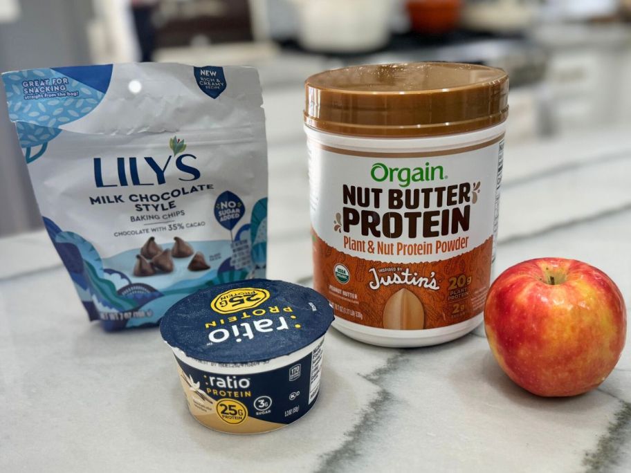 A bag of Lily's Chocolate Chips. protein yogurt, a tub of Orgain Justin's Nut Butter Powder and an Apple