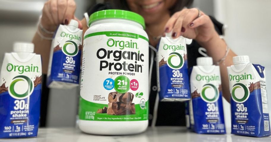 Woman holding Orgain pre-mixed protein shakes next to a tub of Orgain protein powder