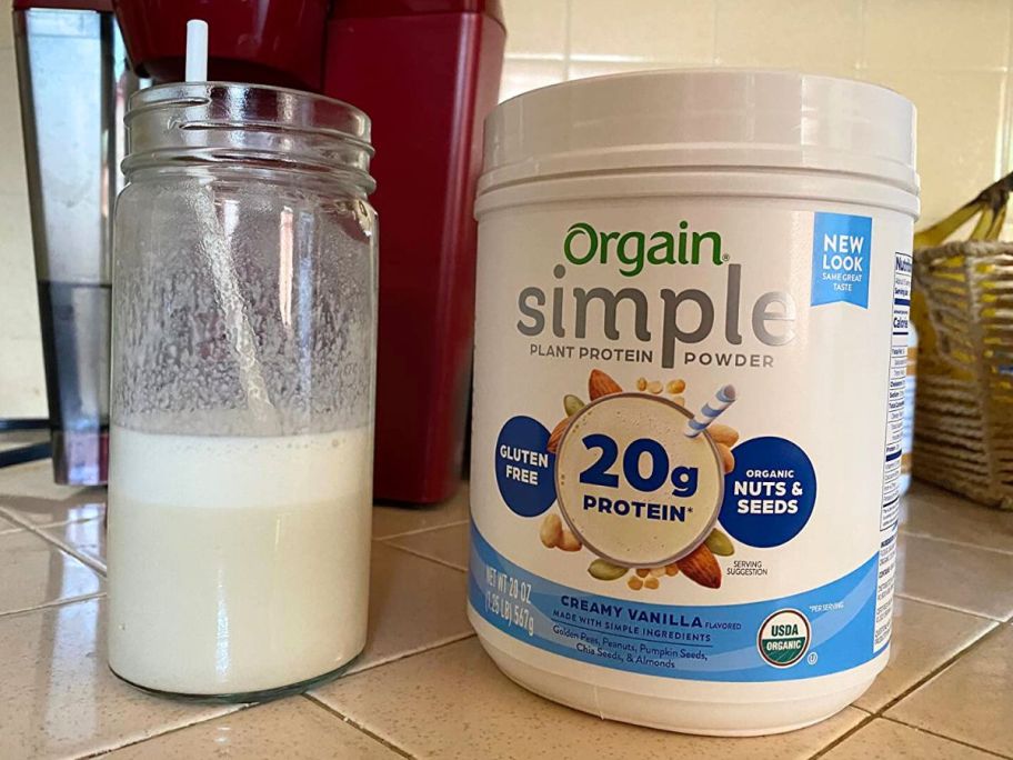 A glass of protein shake and a tub of Orgain Simple Creamy Vanilla Protein Powder