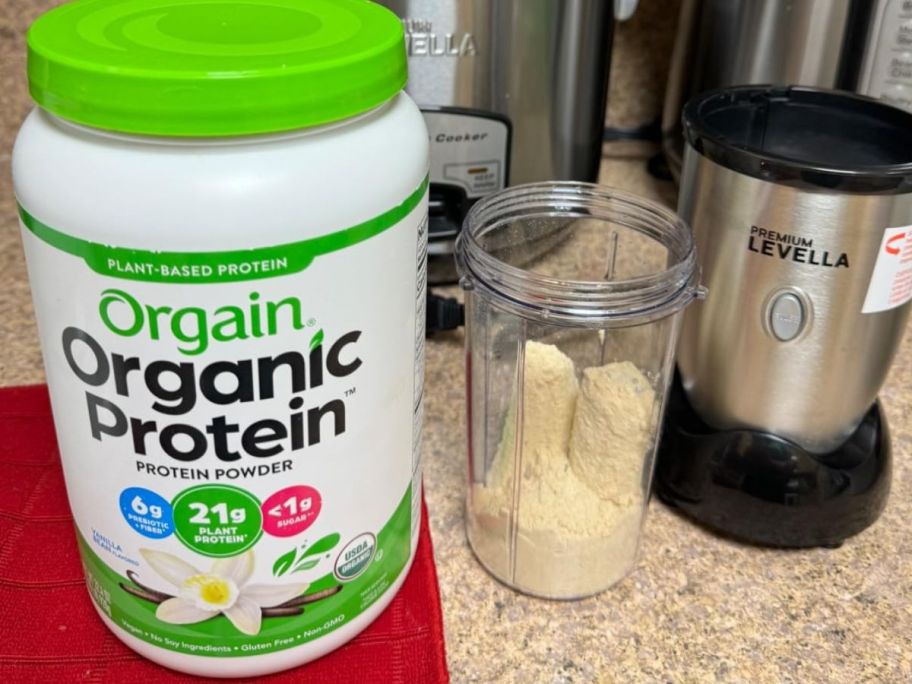 A tub of Orgain Vanilla Bean Protein powder next to a blender cup and blender