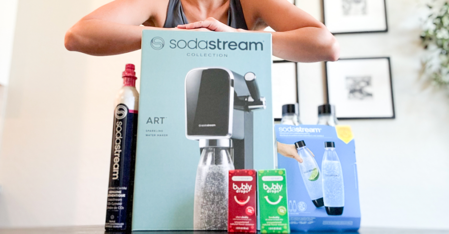 woman standing behind a blue box with a SodaStream machine in it, bubly flavor drops in front and extra SodaStream bottles to the side