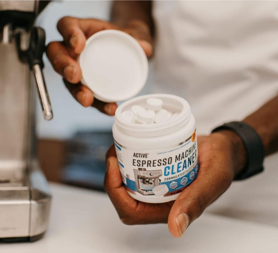 a man holding a jar of espresso cleaner tablets in one hand and the lid in the other