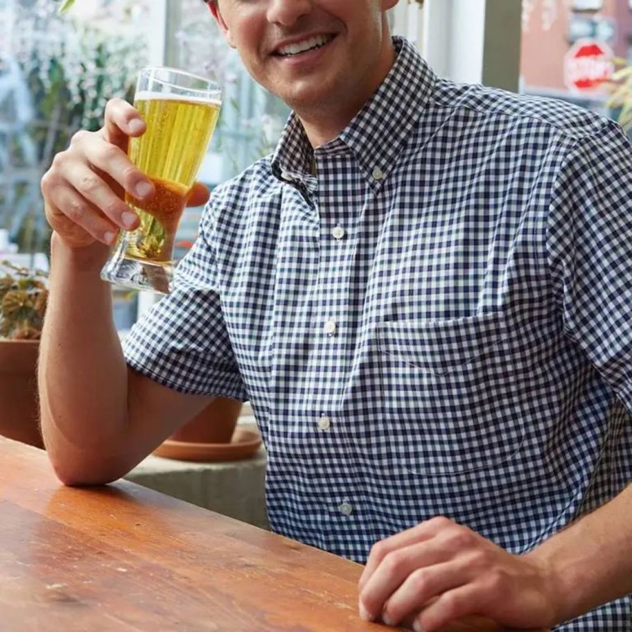 a man in a navy gingham casual button down shirt in a restaurant drinking a draft beer. 