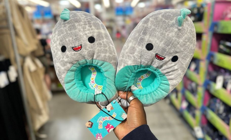 a woman's hand holding a pair of dino squishmallows kids slippers