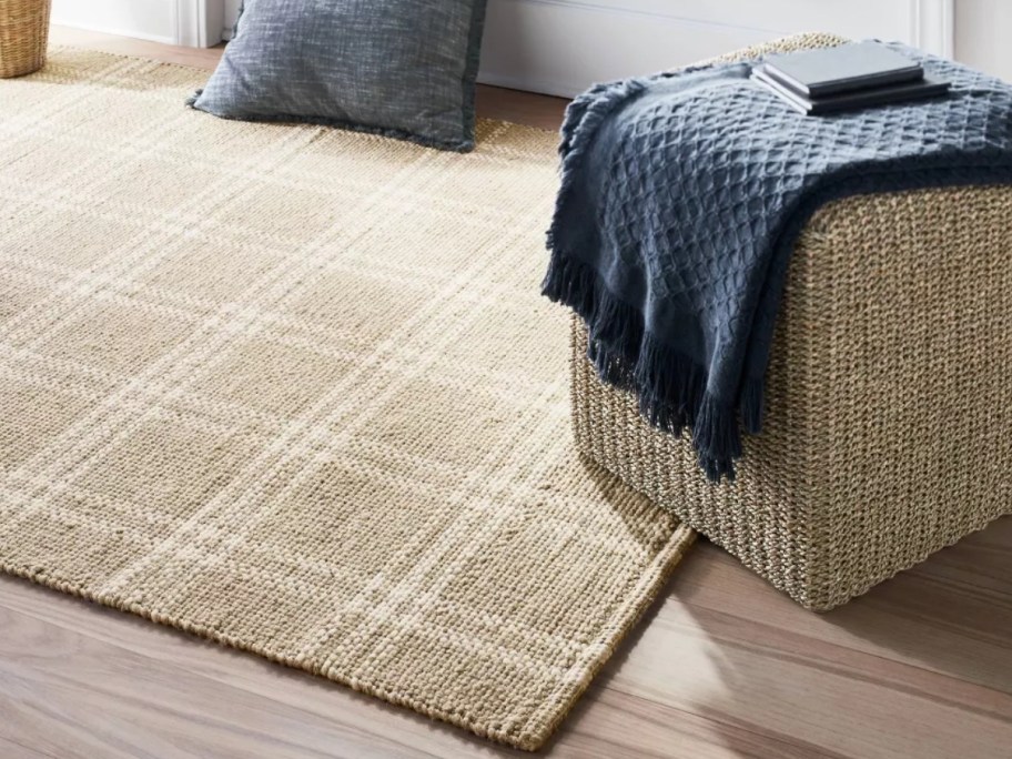 a room with a large tan and white plaid rug and ottoman with a blanket over it