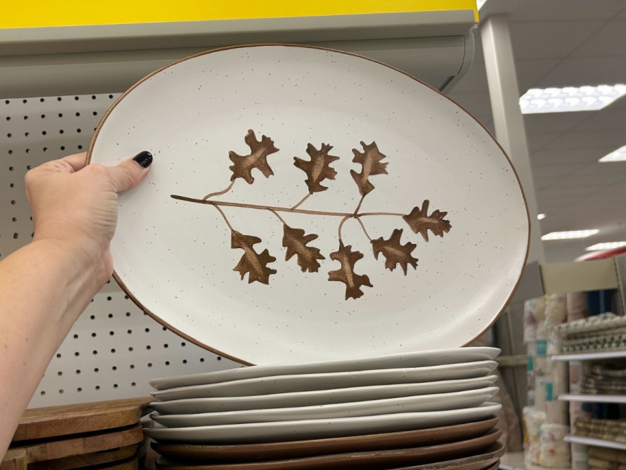 hand holding a large serving plate that is white with brown leaves on it above a stack of more plates on a store shelf