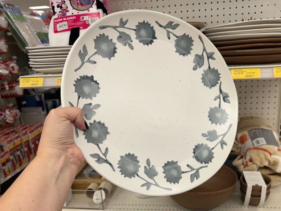 hand holding a large serving plate that's white with blue flowers around the boarder, store shelf with more plates behind it