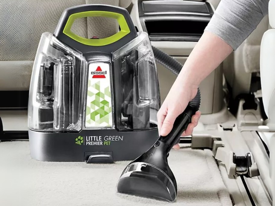 person using a Bissell Little Green Pet Cleaner tool to clean the floor carpet in a vehicle