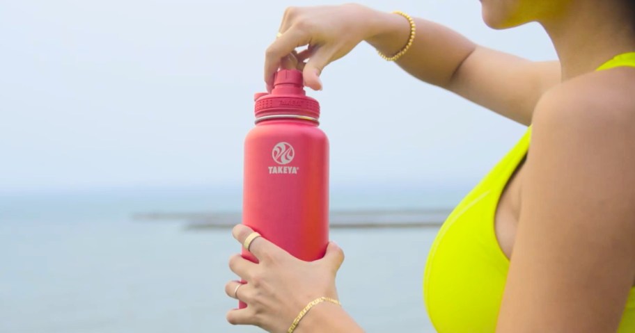 woman wearing a yellow sports bra standing outside by the ocean holding a bright pink water bottle