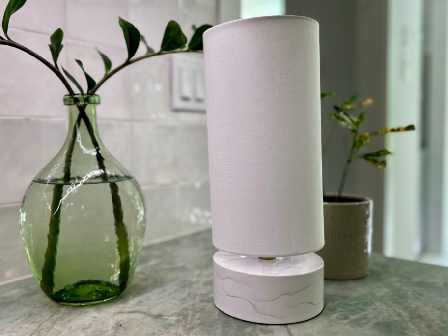 small lamp with a marble base and long white shade sitting on a counter next to a base with leaves in it