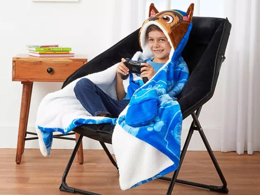 a little boy sitting in a chair with a game controller in his hand, wearing a hooded paw patrol blanket