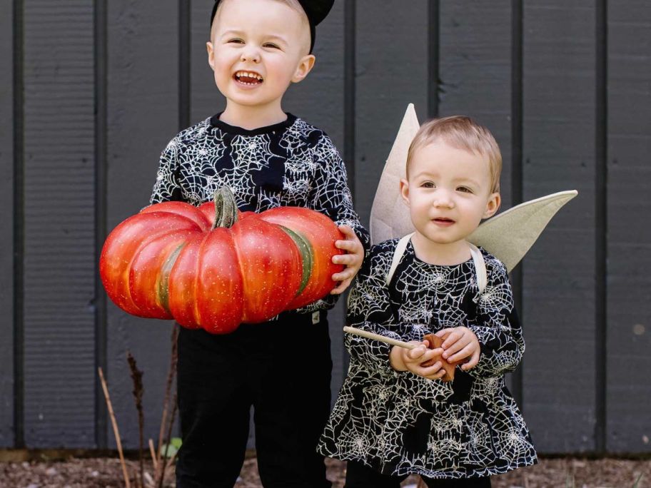 A little boy holding a pumpkin and a little girl wearing angel wings while both wearing Burt's Bees Spiderweb print outfits