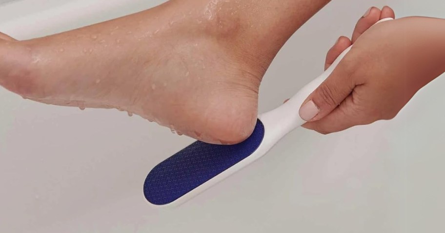 a hand using a blue and white foot file on the heel of a foot in a bathtub to scrub off dead skin