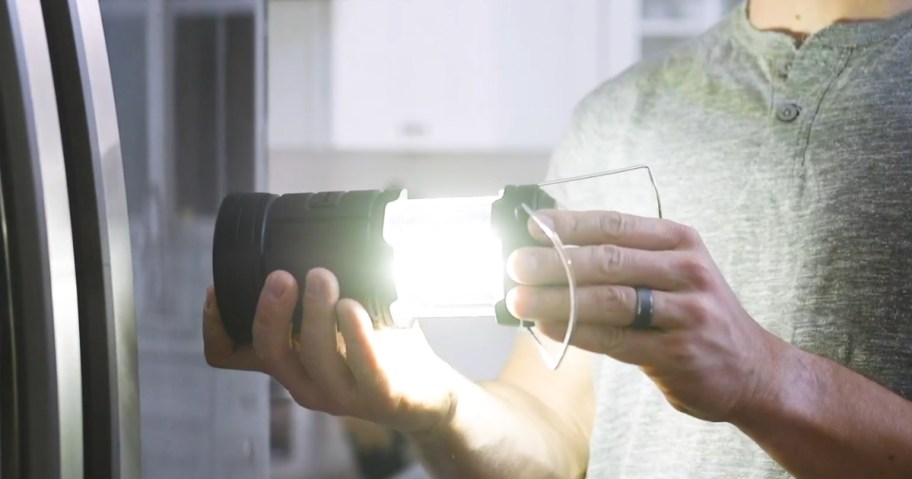 man holding a black camping lantern that's lit up, sideways about to stick it to a stainless steel fridge 