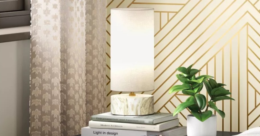 small lamp with a marble base and long white shade sitting on a stack of books next to a faux plant on a beside table