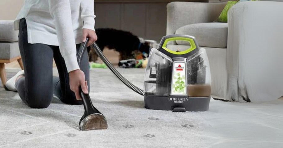 woman using a Bissell Little Green machine to clean up stains off a carpet, there is a dog laying in the background