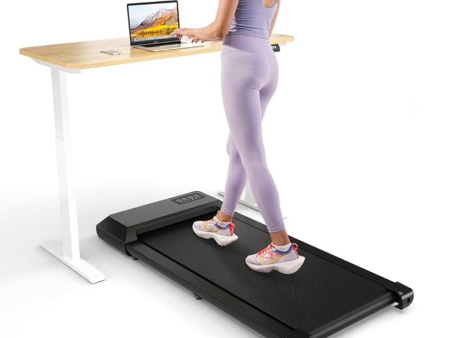 woman in light purple leggings and top walking on a black walking pad at a white and wood top stand up desk working on a laptop