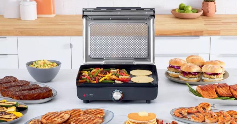 Ninja Sizzle Smokeless Indoor Grill and Griddle on a counter surrounded by loads of food 