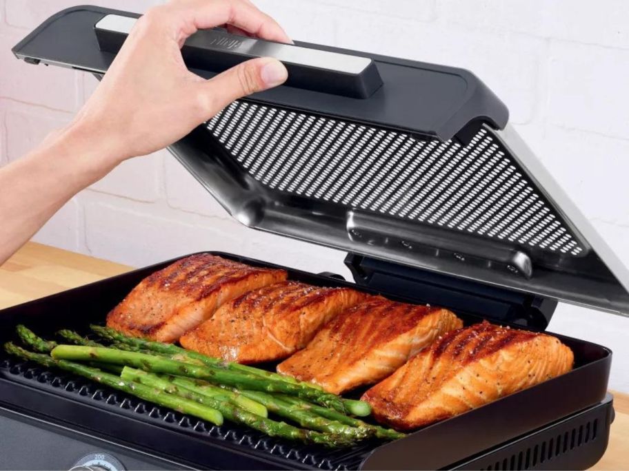 A person opening the lid on a Ninja Sizzle Smokeless Indoor Grill and Griddle to reveal salmon and asparagus