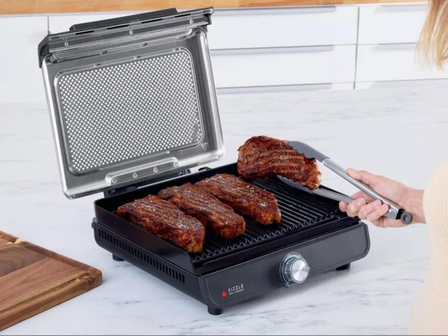 A person turning steaks on a Ninja Sizzle Smokeless Indoor Grill and Griddle