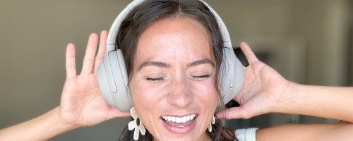 woman with brown hair wearing silver Sony headphones with her hands on them, her eyes shut and smiling