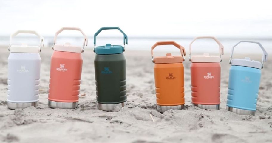 6 Stanley Iceflow Jugs in different colors set in the sand on a beach