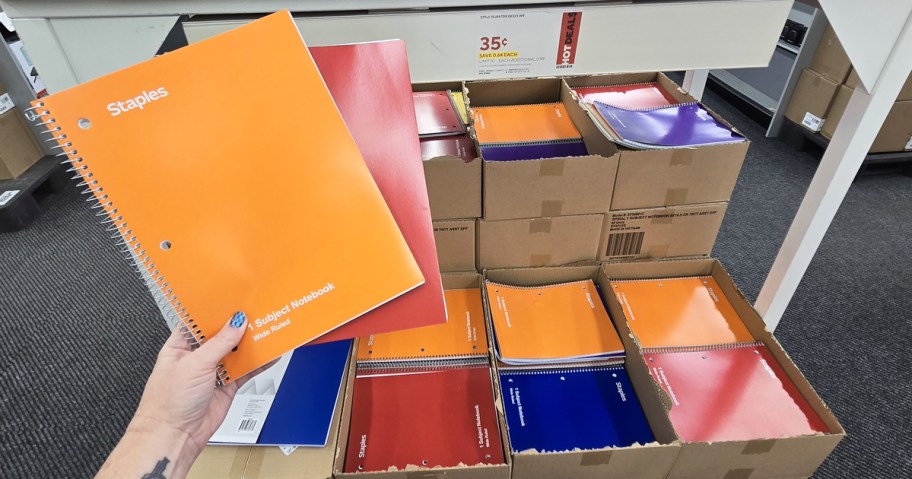 hand holding up orange and red 1-subject notebooks in front of cases of more notebooks