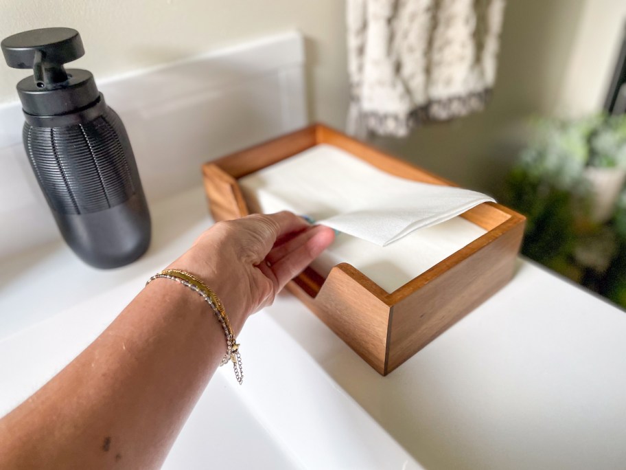 hand taking white disposable hand towel napkin out of wood holder on bathroom sink