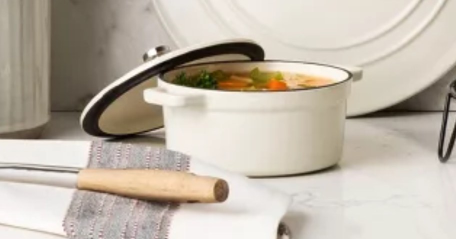 white mini dutch oven with the lid resting on the side of it, large spoon and kitchen towel in front