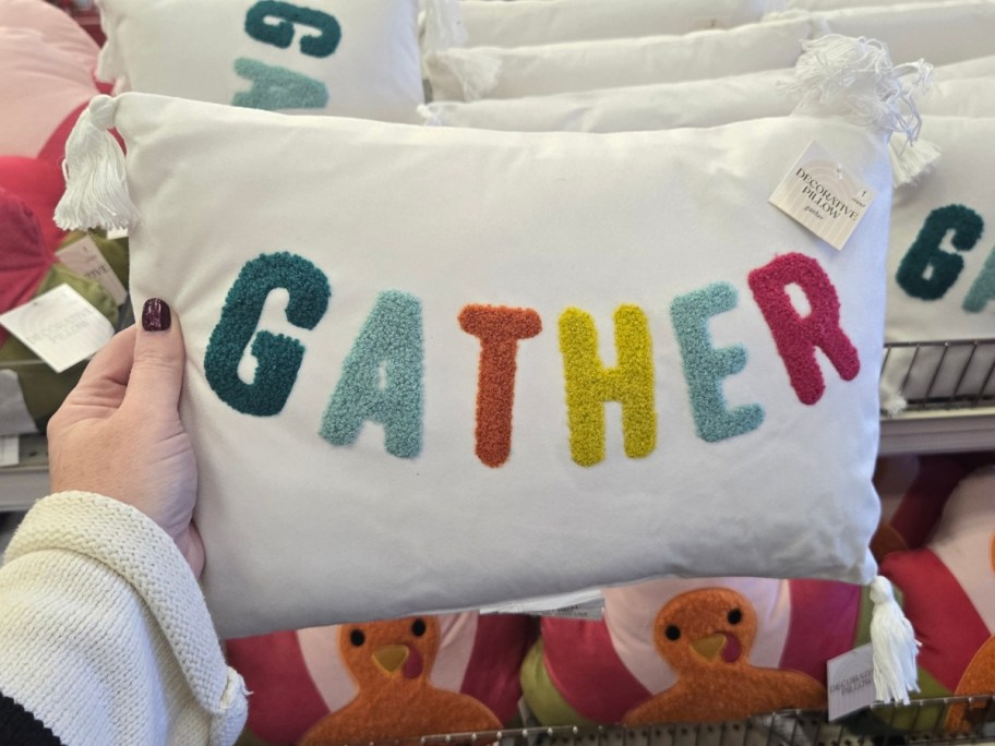 hand holding a white rectangle throw pillow with the word "Gather" in different colors for each letter