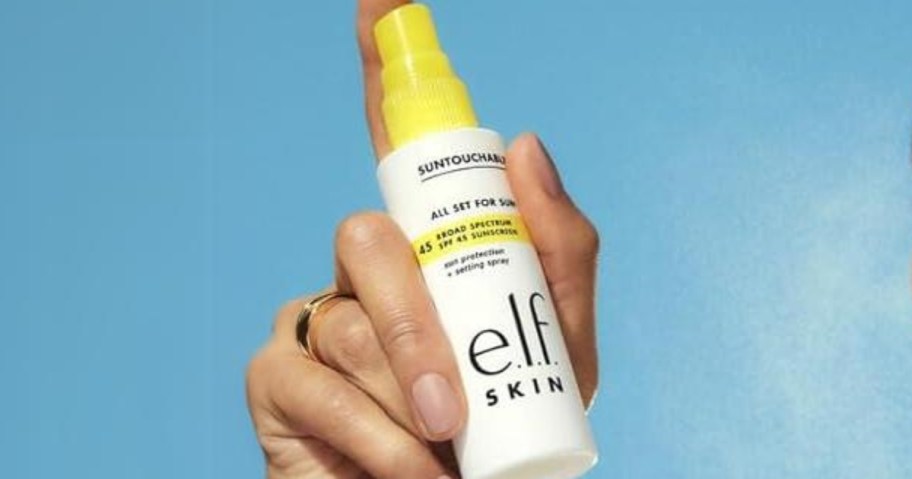 hand holding a white and yellow bottle of e.l.f. Skin sunscreen spray, blue cloudy sky behind it