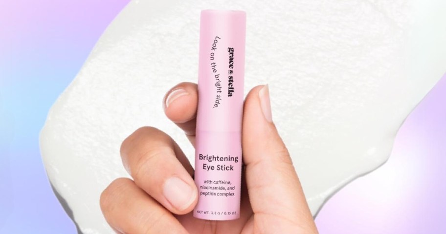 hand holding a pink tube of Grace Stella Brightening Under Eye Stick, the balm spread out behind it on a multi color background