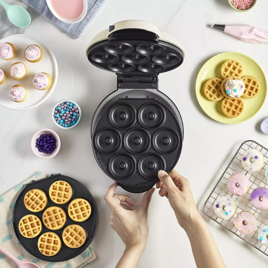 person reaching for a Dash Treat MultiMaker, plate with mini waffles to one size, rack with mini donuts cooling to the other site and plate of mini cupcakes and more mini waffles on each side