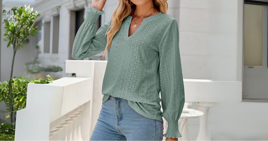 woman wearing a light sage green long sleeve blouse with eyelet details standing outside with trees and a white stone railing and building behind her