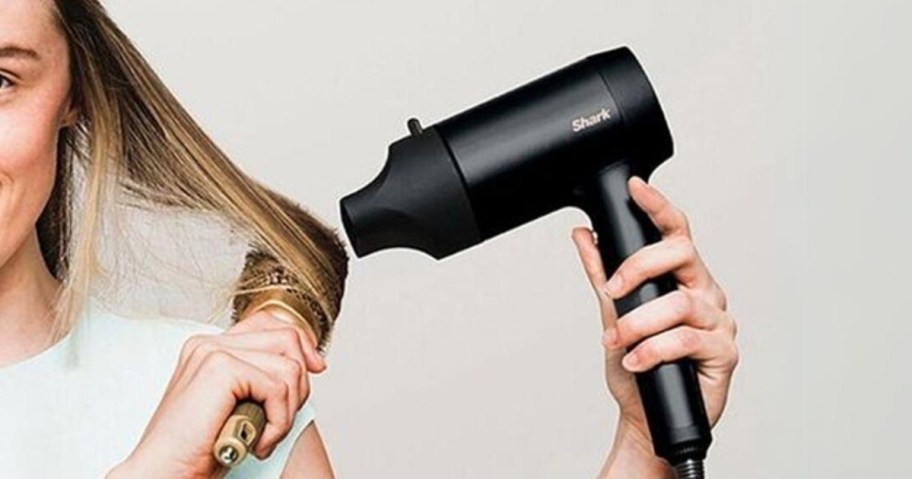 woman holding a black Shark hairdryer with an attachment on it, using a round brush to dry and style her hair