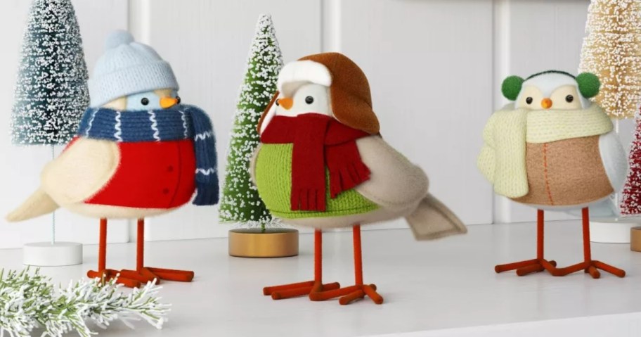 3 fabric birds in winter clothing on a mantle with Christmas decor and bristle brush trees behind them