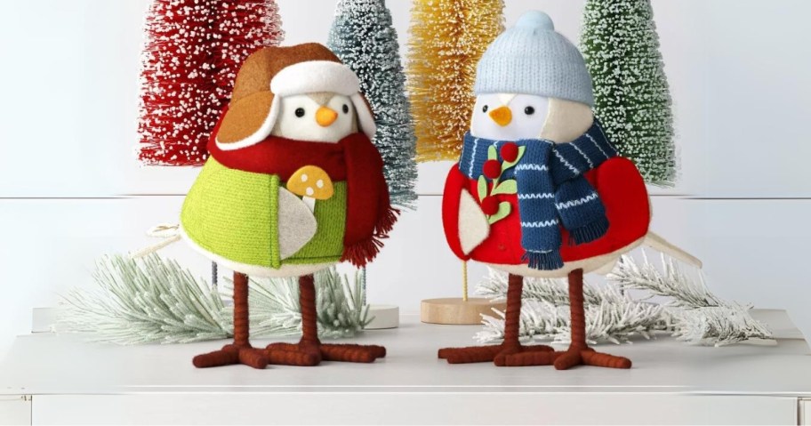 2 fabric birds in winter clothing, one holding a mushroom, one holding a holly leaf and berry stem, bristle brush Christmas trees behind them