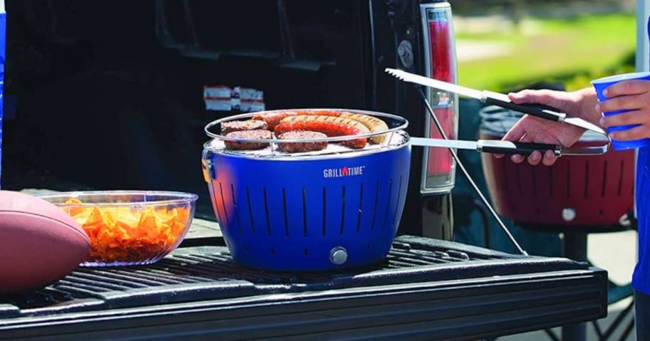 a person grilling food on a blue tabletop portable grill on the back of a pickup truck in a parking lot