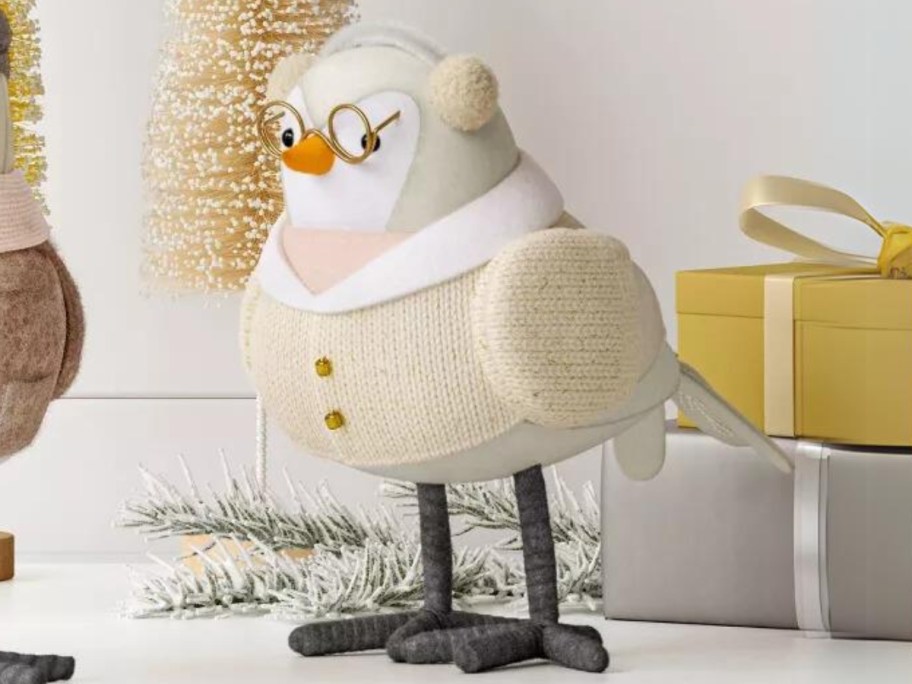 fabric holiday bird in cream sweater with white collar, glasses and ear muffs, presents and bristle brush trees behind her