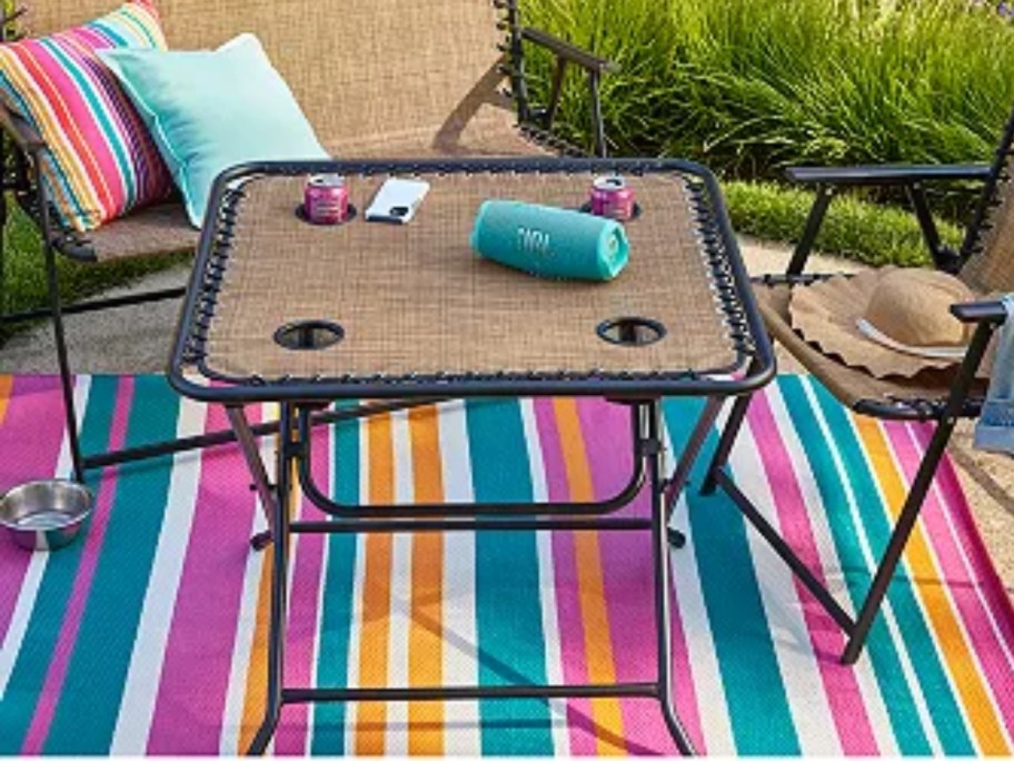 a brown folding patio table with cup holders on a patio next to chairs on top of a multi color stripe rug