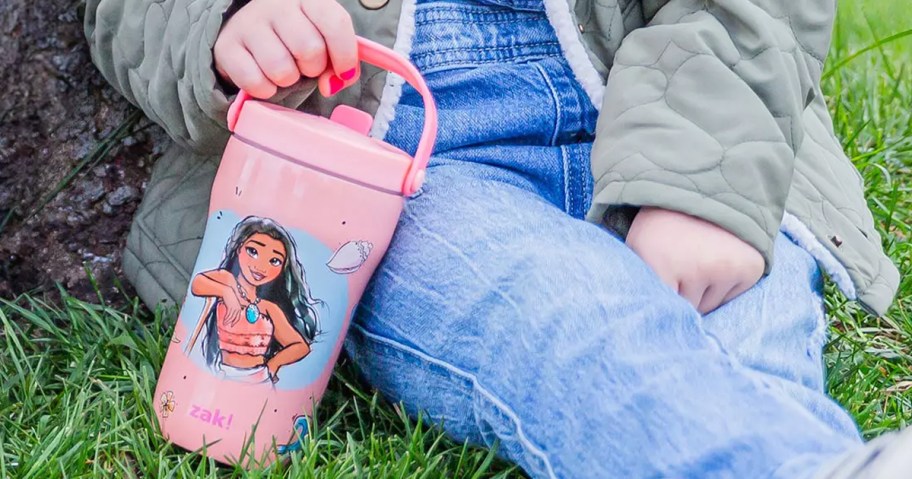 little girl sitting on the ground holding a pink Disney Moana tumbler