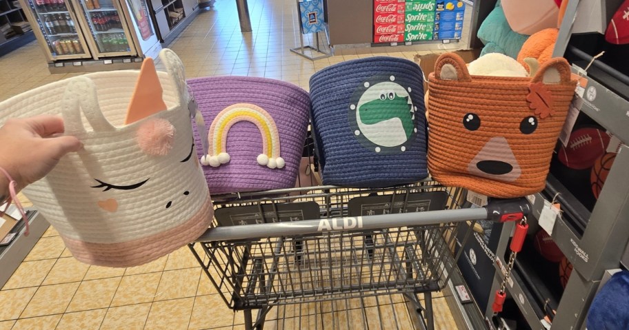 hand holding a white and pink cat rope basket, more behind in the cart in purple with rainbow, blue with dinosaur, orange with bear