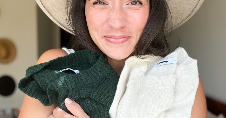 woman with a tan hat smiling at the camera holding a green and an off white sweater