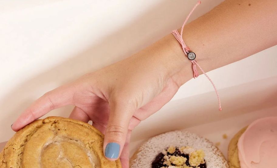 a womans hand and wrist shown wearing a pink multistrand bracelet and reaching into a cookie box to grab a cookie