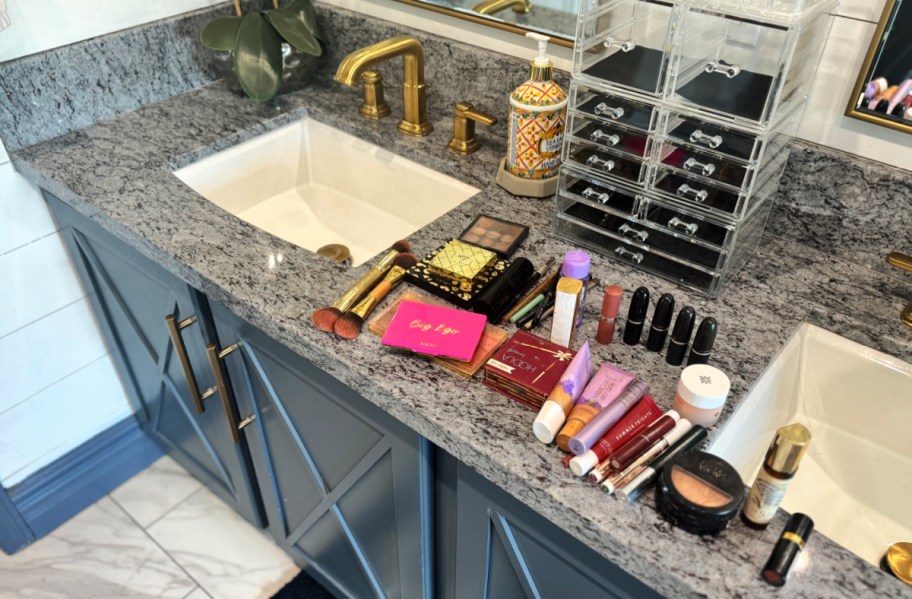 many cosmetic items laid out on a bathroom counter in front of an empty sorbus makeup organizer, demonstrating how much it will hold