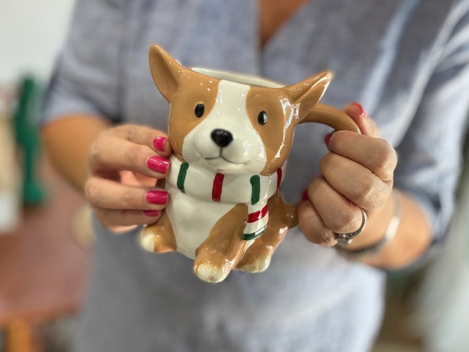 woman's hands holding a coffee mug shaped like a tan and white Corgi dog with a red, white, and green scarf