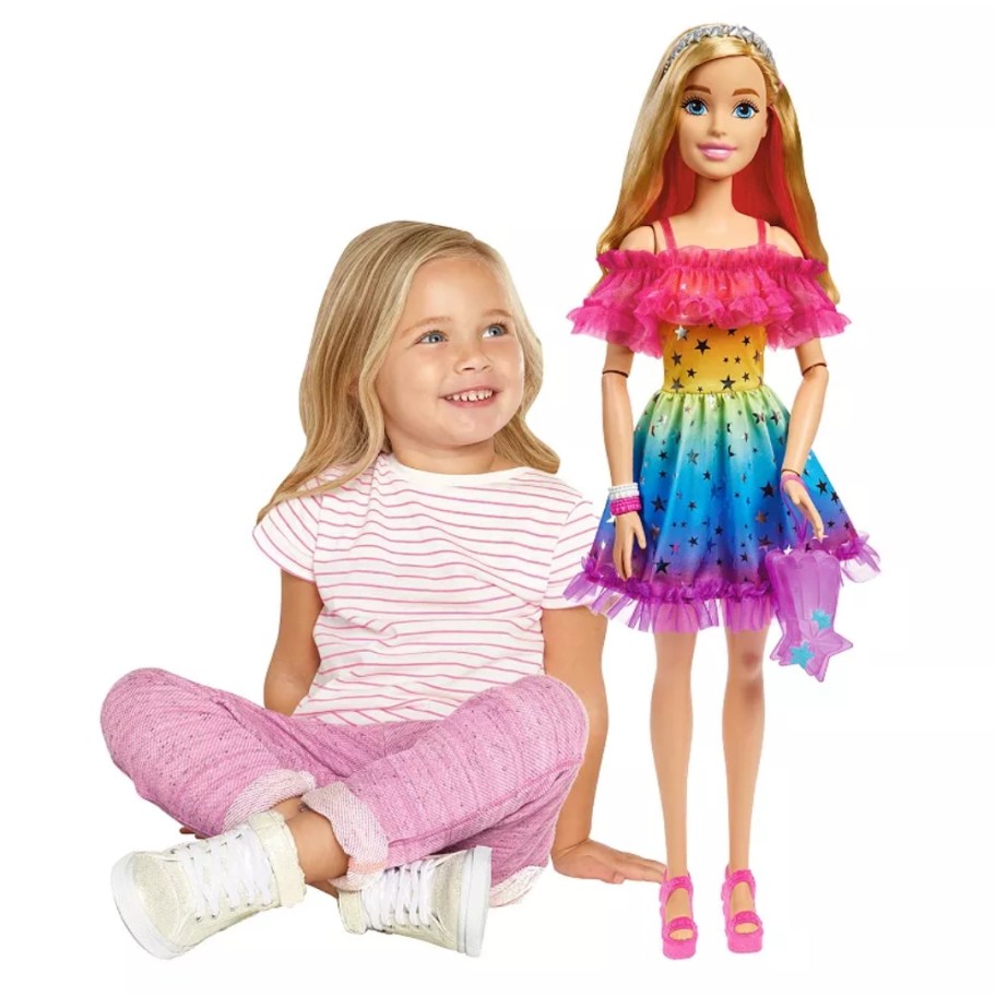 a little girl sitting next to a large Barbie doll wearing a colorful dress