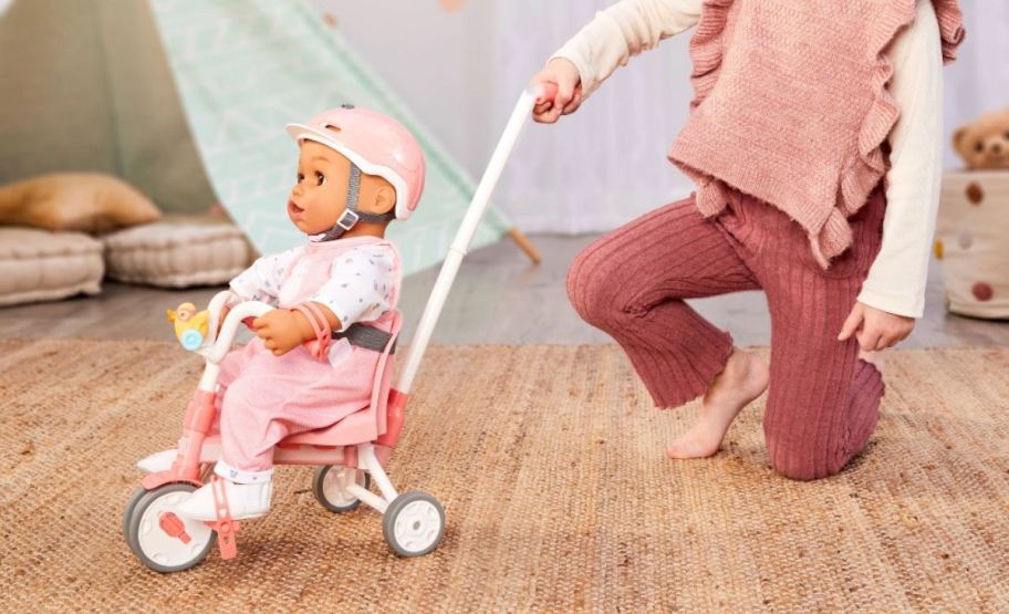 a little girl pushing a baby doll on a tricycle