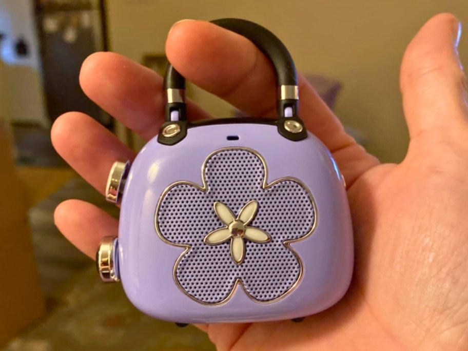 hand holding a mini purple and black Bluetooth speaker that looks like a purse and has a flower shape on it